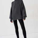 A woman modeling an oversized dark gray turtleneck sweater, paired with black leggings and black-and-white striped kitten heels, against a plain white studio background.