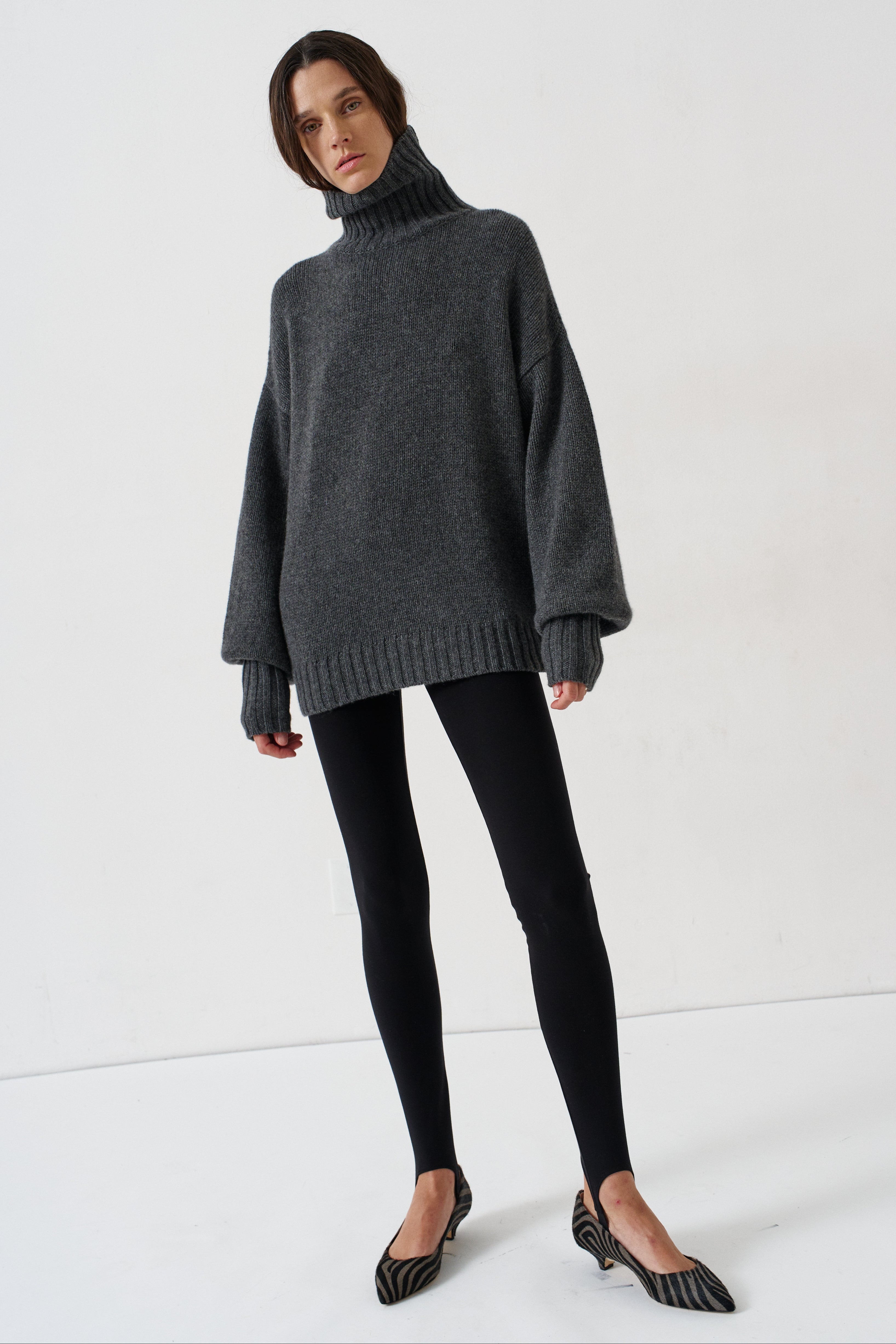 A woman modeling an oversized dark gray turtleneck sweater, paired with black leggings and black-and-white striped kitten heels, against a plain white studio background.
