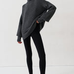 A woman modeling an oversized dark gray turtleneck sweater, paired with black leggings and black-and-white striped kitten heel flats, against a plain white studio background.