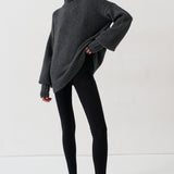 A woman modeling an oversized dark gray turtleneck sweater, paired with black leggings and black-and-white striped kitten heel flats, against a plain white studio background.
