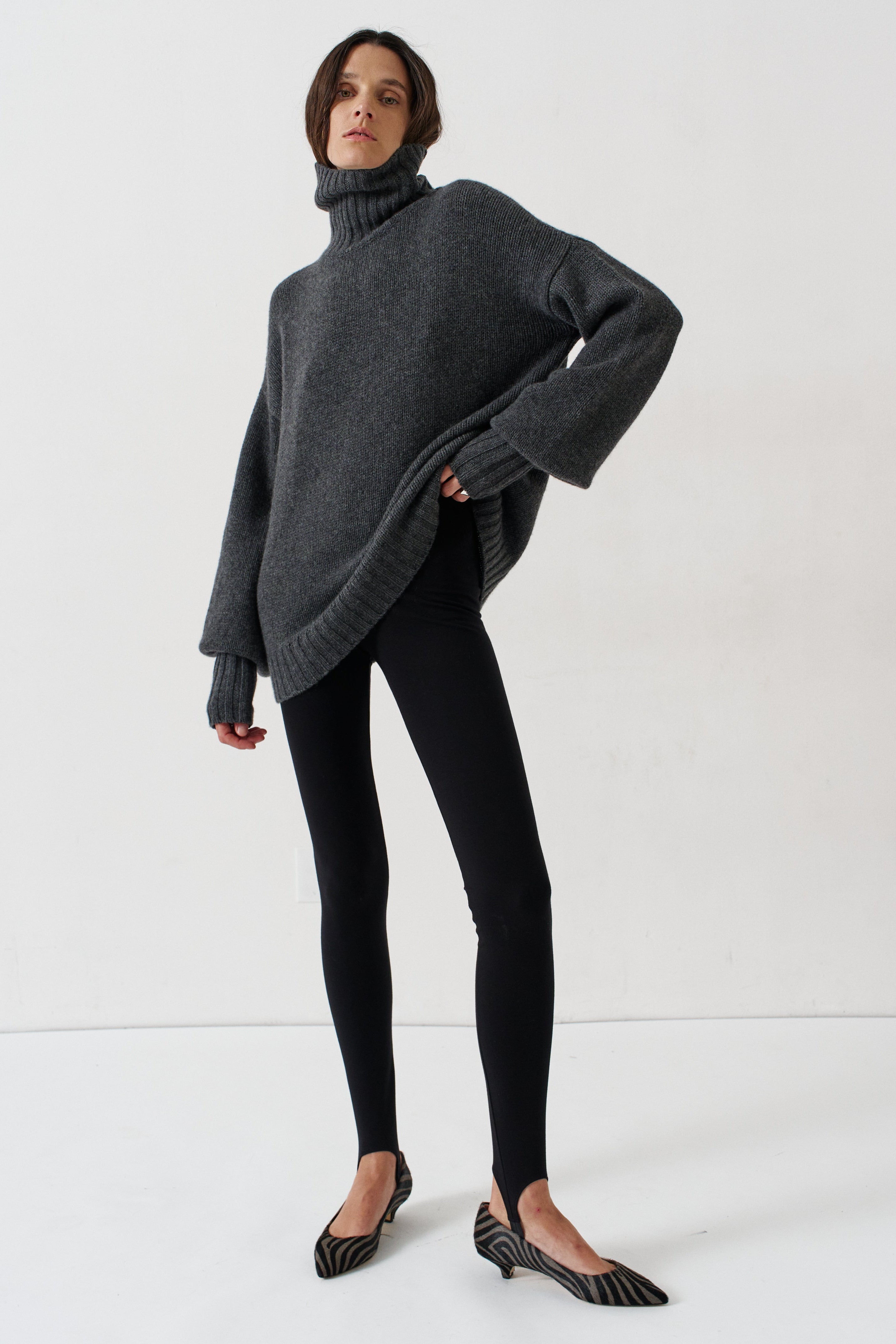 A woman modeling an oversized dark gray turtleneck sweater, paired with black leggings and black-and-white striped kitten heel flats, against a plain white studio background.