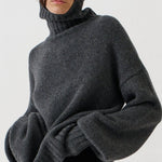 A model wearing a dark gray oversized knit turtleneck sweater, black leggings, and black-and-white striped kitten heel flats, standing against a plain white studio background.