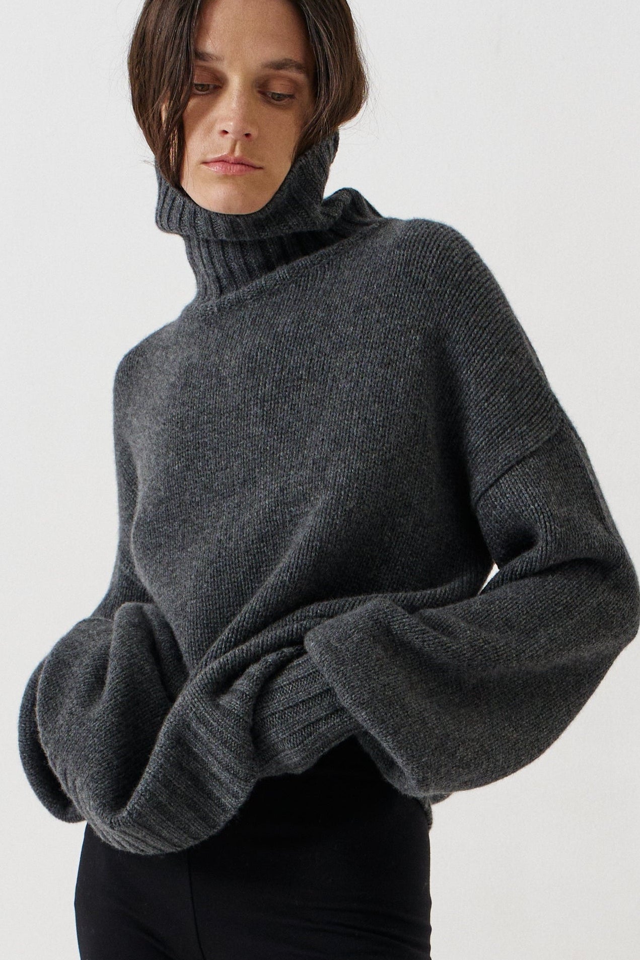 A model wearing a dark gray oversized knit turtleneck sweater, black leggings, and black-and-white striped kitten heel flats, standing against a plain white studio background.