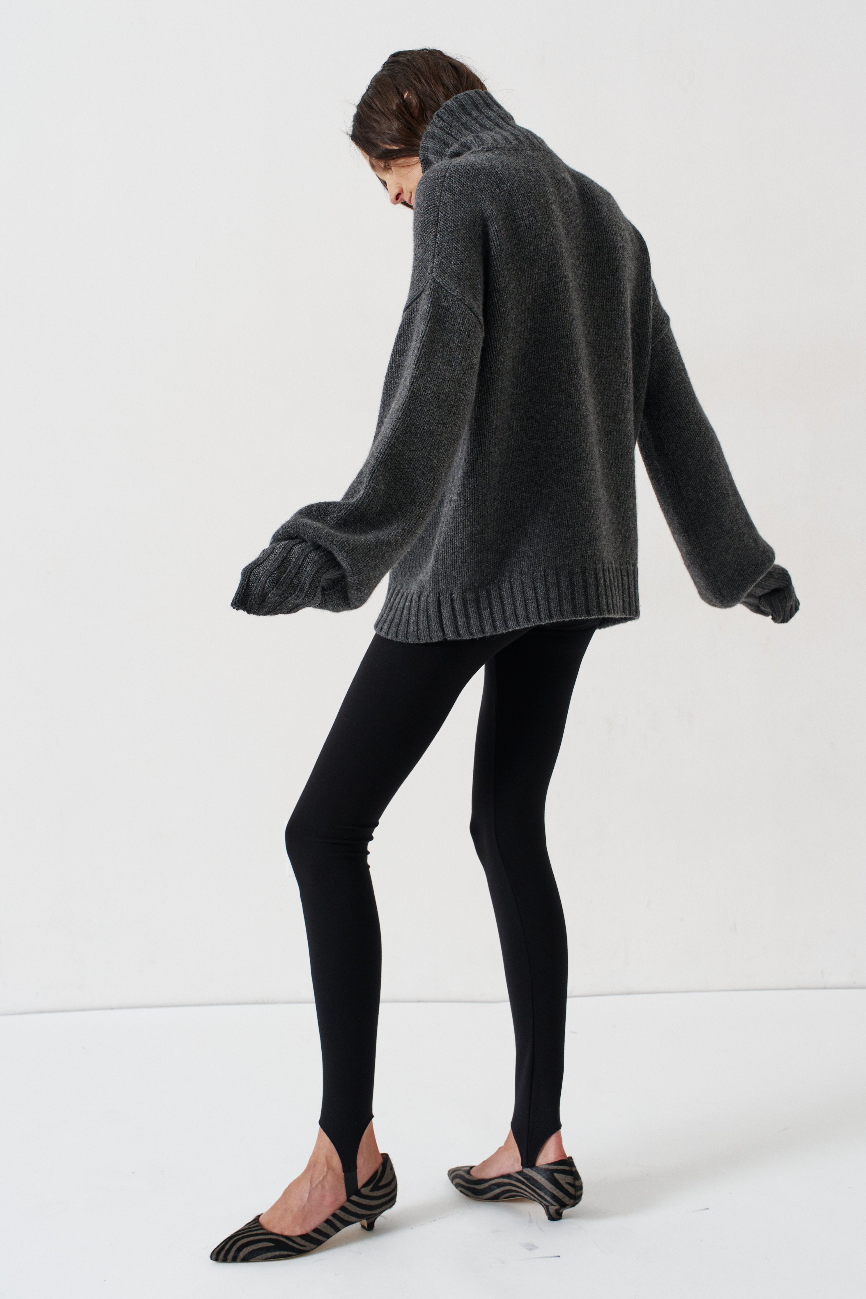 A model wearing a dark gray oversized knit sweater, black leggings, and black pointed-toe slingback heels, captured from a side-back angle against a clean white studio background.