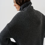 A person wearing a dark charcoal gray ribbed knit turtleneck sweater, shown from a side-back angle against a plain light background.