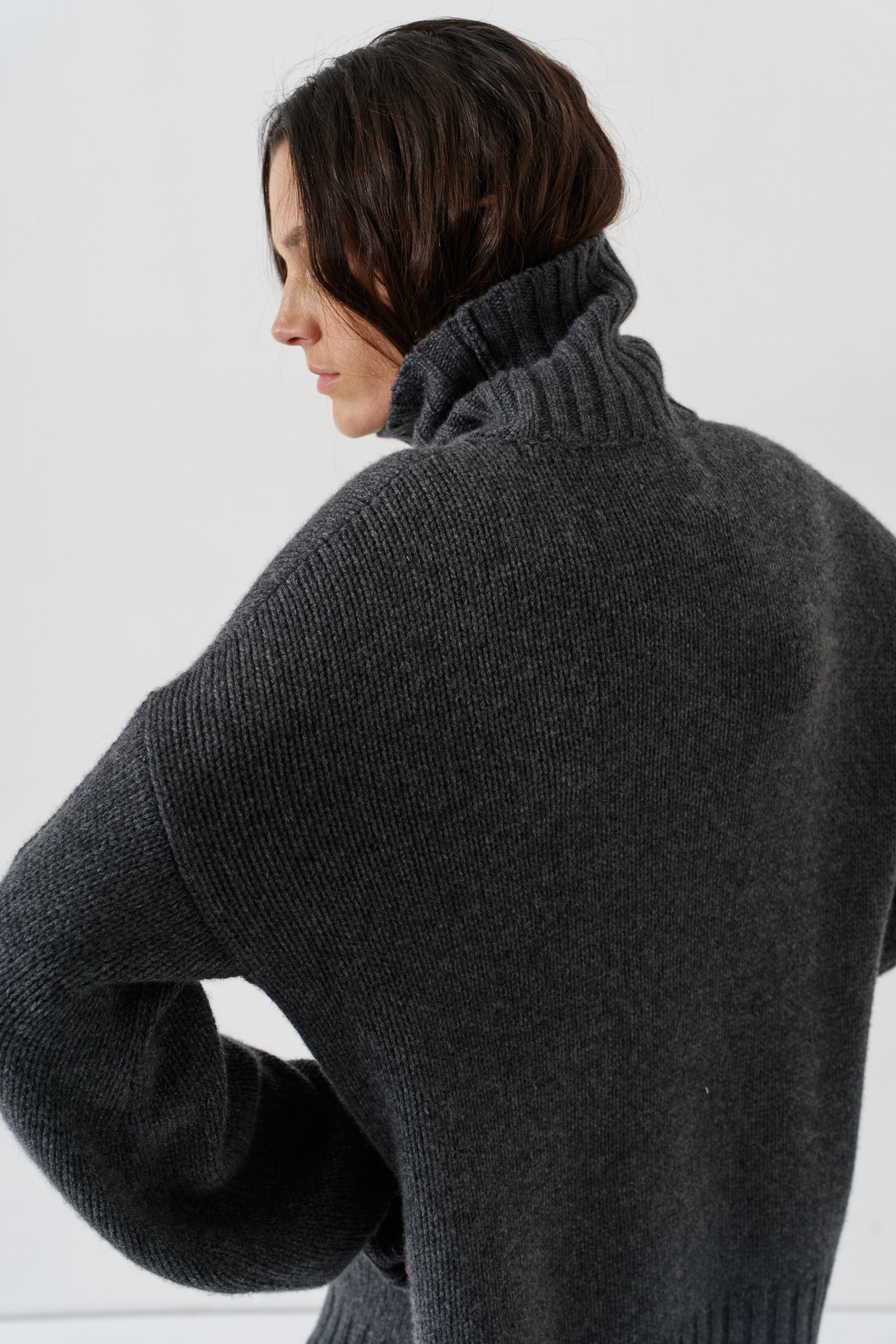 A person wearing a dark charcoal gray ribbed knit turtleneck sweater, shown from a side-back angle against a plain light background.
