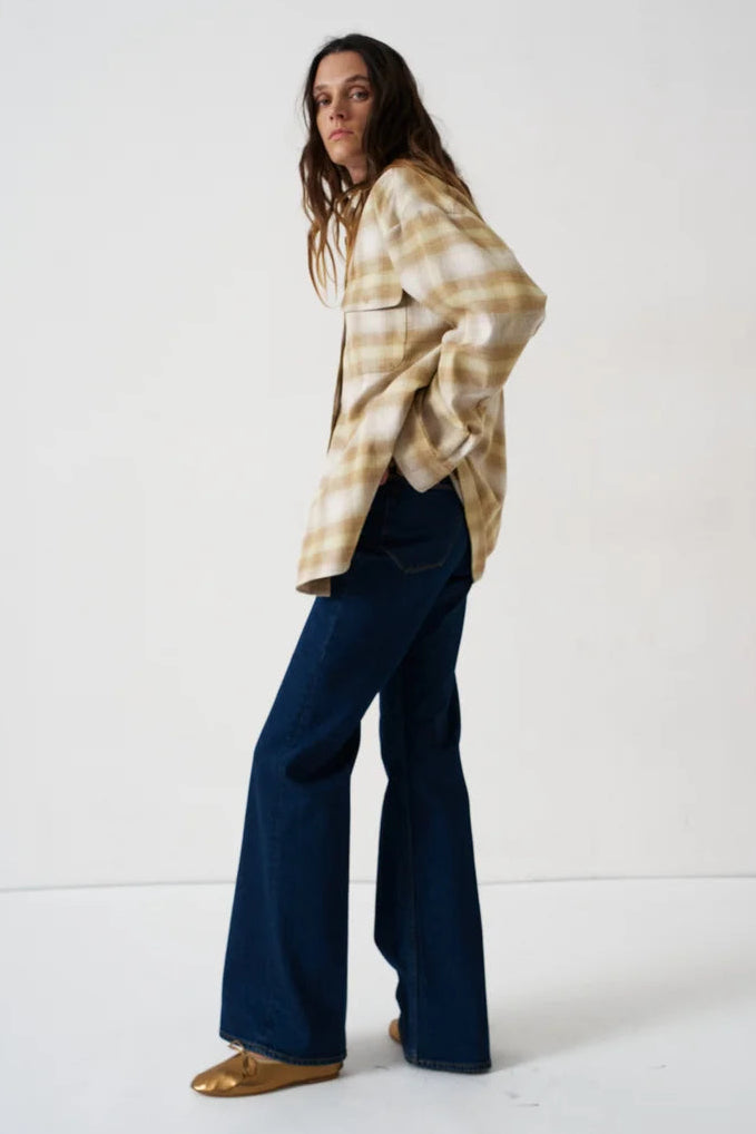 A woman modeling a beige and brown plaid flannel shirt with dark blue wide-leg jeans and gold flat shoes, standing against a plain white studio background.