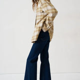 A woman modeling a beige and brown plaid flannel shirt with dark blue wide-leg jeans and gold flat shoes, standing against a plain white studio background.