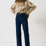 A woman stands against a plain white studio background wearing a cropped beige-and-brown plaid flannel shirt, dark blue wide-leg jeans, and brown shoes.