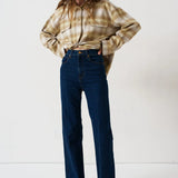 A woman stands against a plain white studio background wearing a cropped beige-and-brown plaid flannel shirt, dark blue wide-leg jeans, and brown shoes.