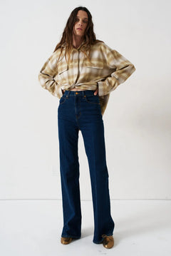A woman stands against a plain white studio background wearing a cropped beige-and-brown plaid flannel shirt, dark blue wide-leg jeans, and brown shoes.