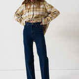 A woman modeling a beige and brown plaid button-up shirt tucked into dark blue high-waisted straight-leg jeans, standing against a plain white studio background.