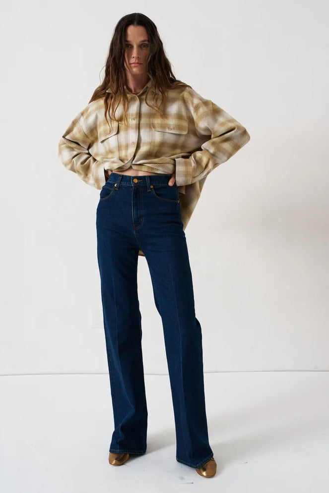 A woman modeling a beige and brown plaid button-up shirt tucked into dark blue high-waisted straight-leg jeans, standing against a plain white studio background.