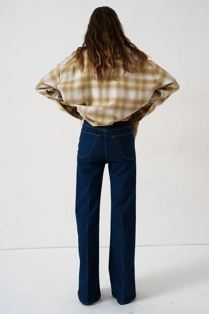 A woman is shown from behind wearing a beige and brown plaid flannel shirt and dark blue wide-leg jeans, standing against a plain white studio background.