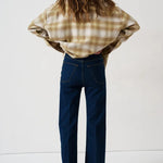 A woman is shown from behind wearing a beige and brown plaid flannel shirt and dark blue wide-leg jeans, standing against a plain white studio background.