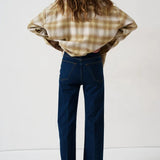A woman is shown from behind wearing a beige and brown plaid flannel shirt and dark blue wide-leg jeans, standing against a plain white studio background.