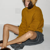 A model sits on a white floor against a white wall, wearing a mustard yellow knit sweater, a brown fur-trimmed hat, and brown fur slippers.