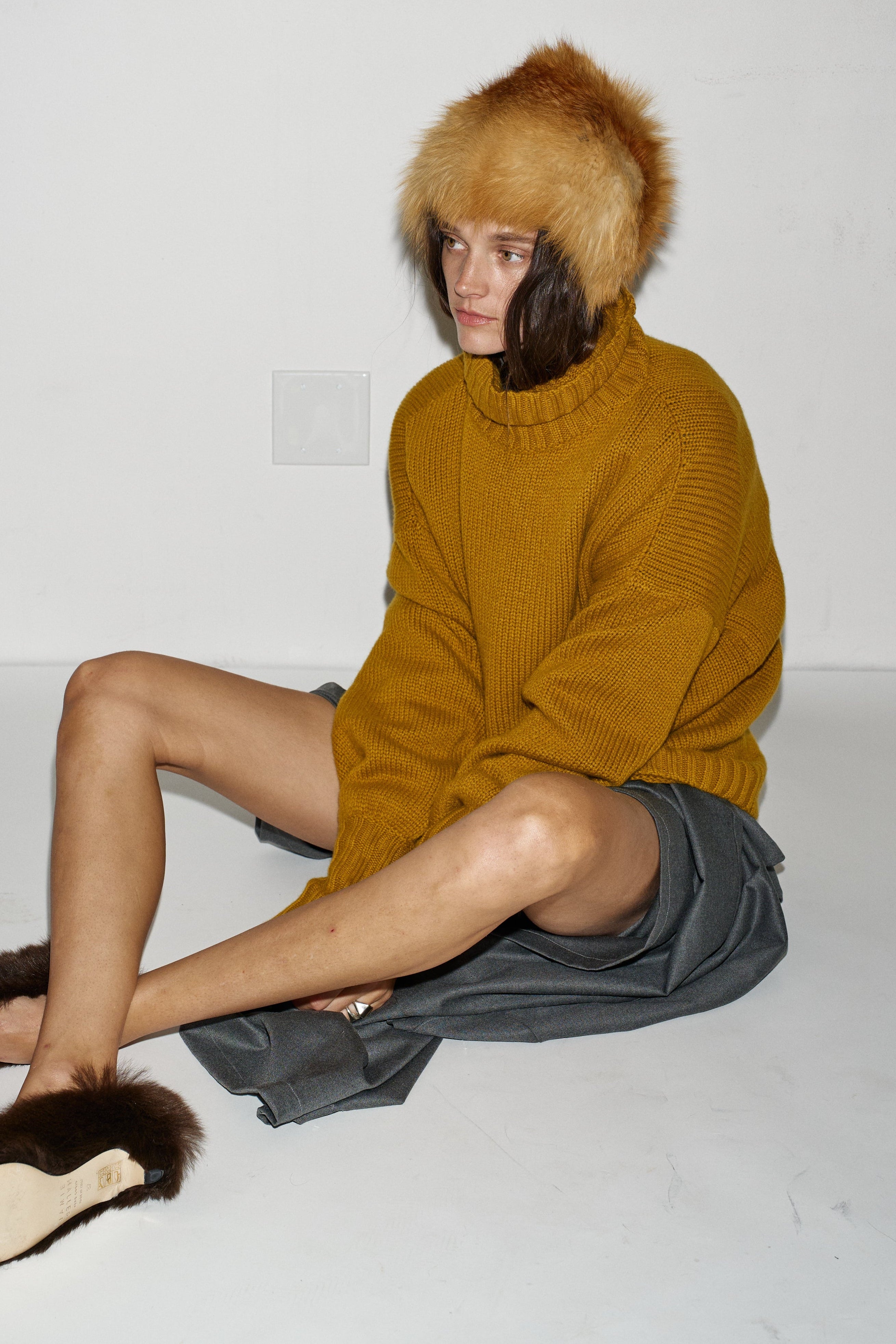 A model sits on a white floor against a white wall, wearing a mustard yellow knit sweater, a brown fur-trimmed hat, and brown fur slippers.
