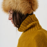 A close-up side profile of a person wearing a large tan and brown fur hat with a ribbed mustard yellow turtleneck sweater against a plain light gray background.