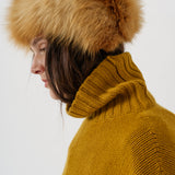 A close-up side profile of a person wearing a large tan and brown fur hat with a ribbed mustard yellow turtleneck sweater against a plain light gray background.