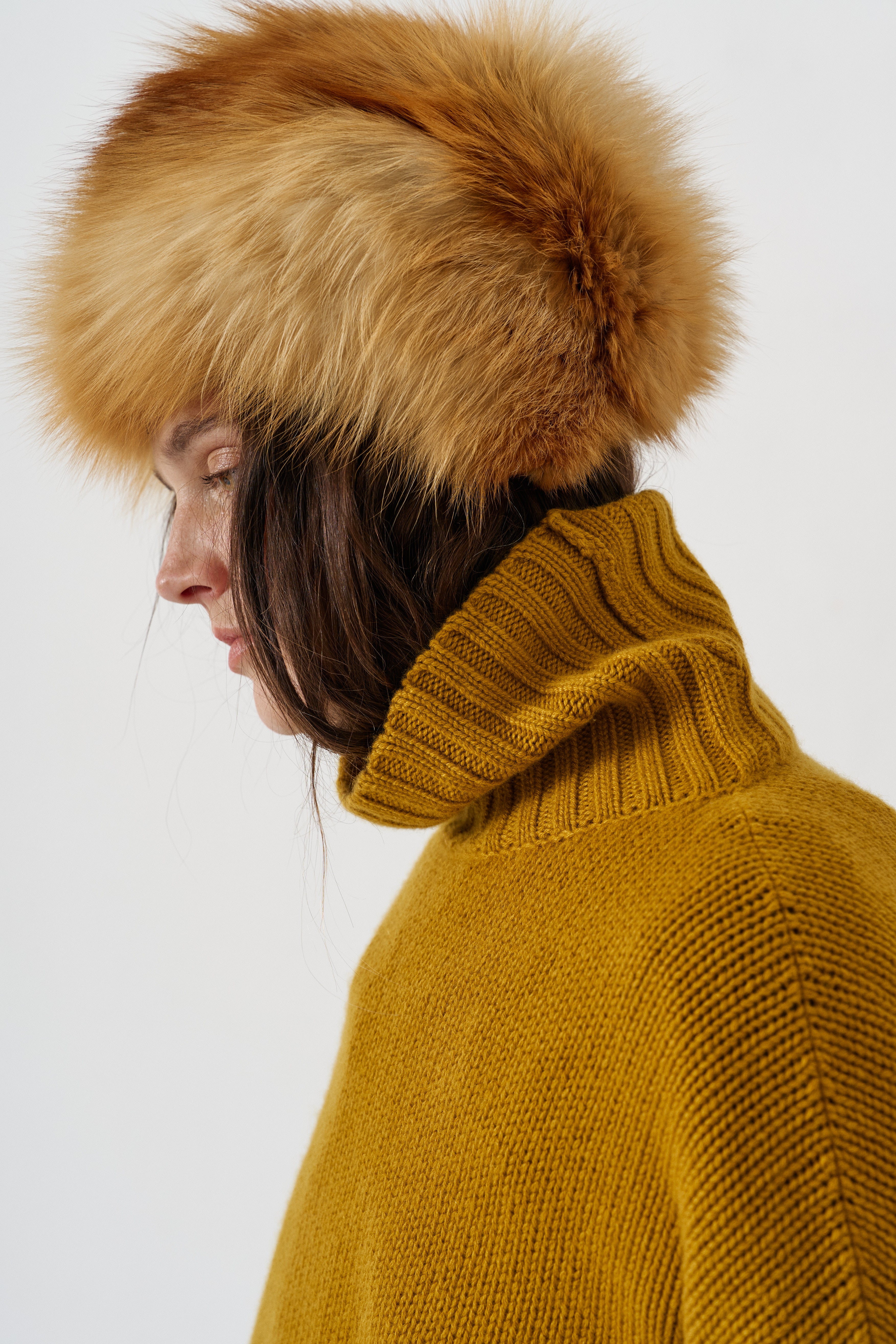 A close-up side profile of a person wearing a large tan and brown fur hat with a ribbed mustard yellow turtleneck sweater against a plain light gray background.