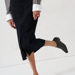 A person wearing a black midi skirt with a front slit, paired with a striped shirt and black ankle boots, captured in a studio setting.