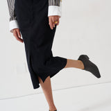 A person wearing a black midi skirt with a front slit, paired with a striped shirt and black ankle boots, captured in a studio setting.