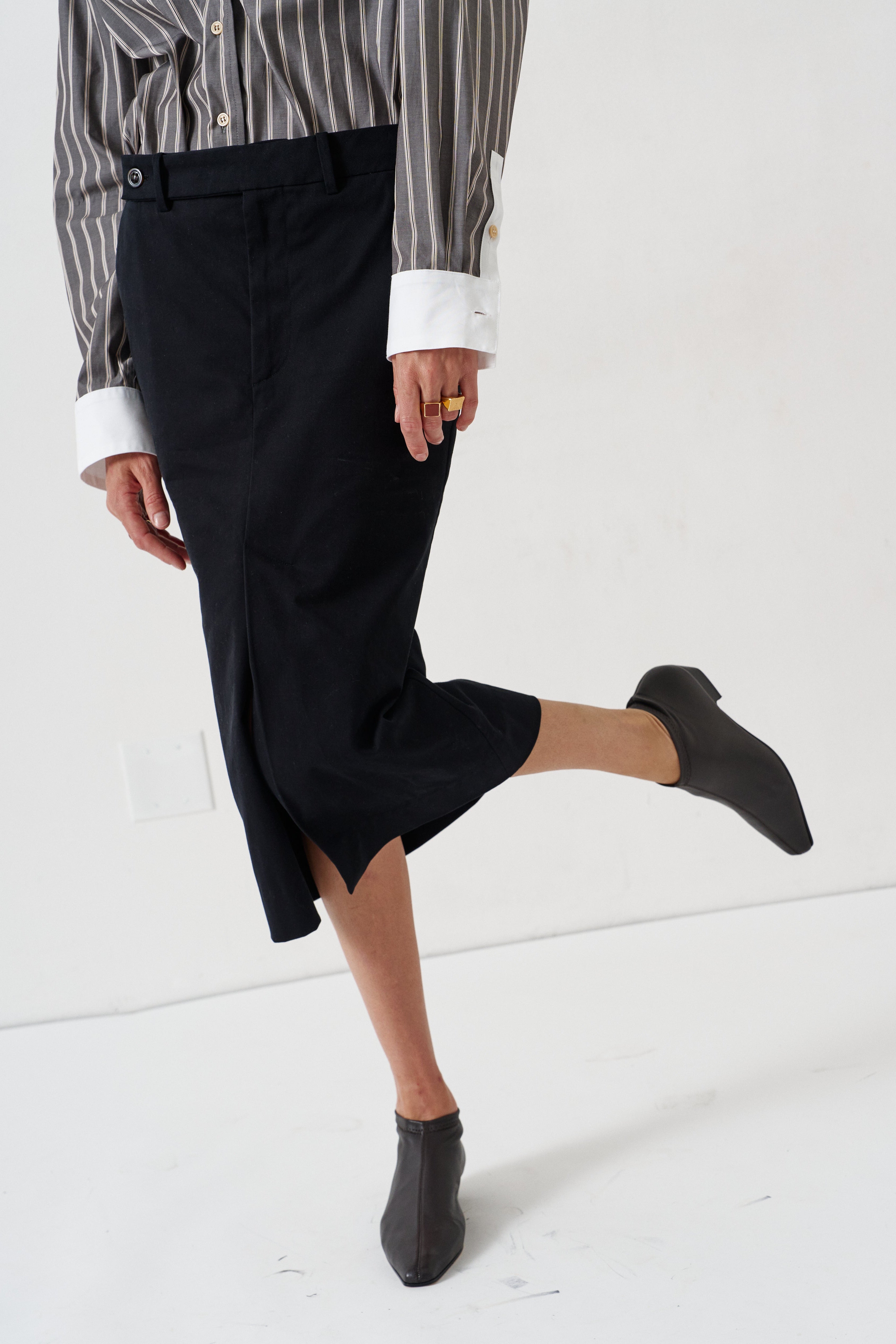 A person wearing a black midi skirt with a front slit, paired with a striped shirt and black ankle boots, captured in a studio setting.