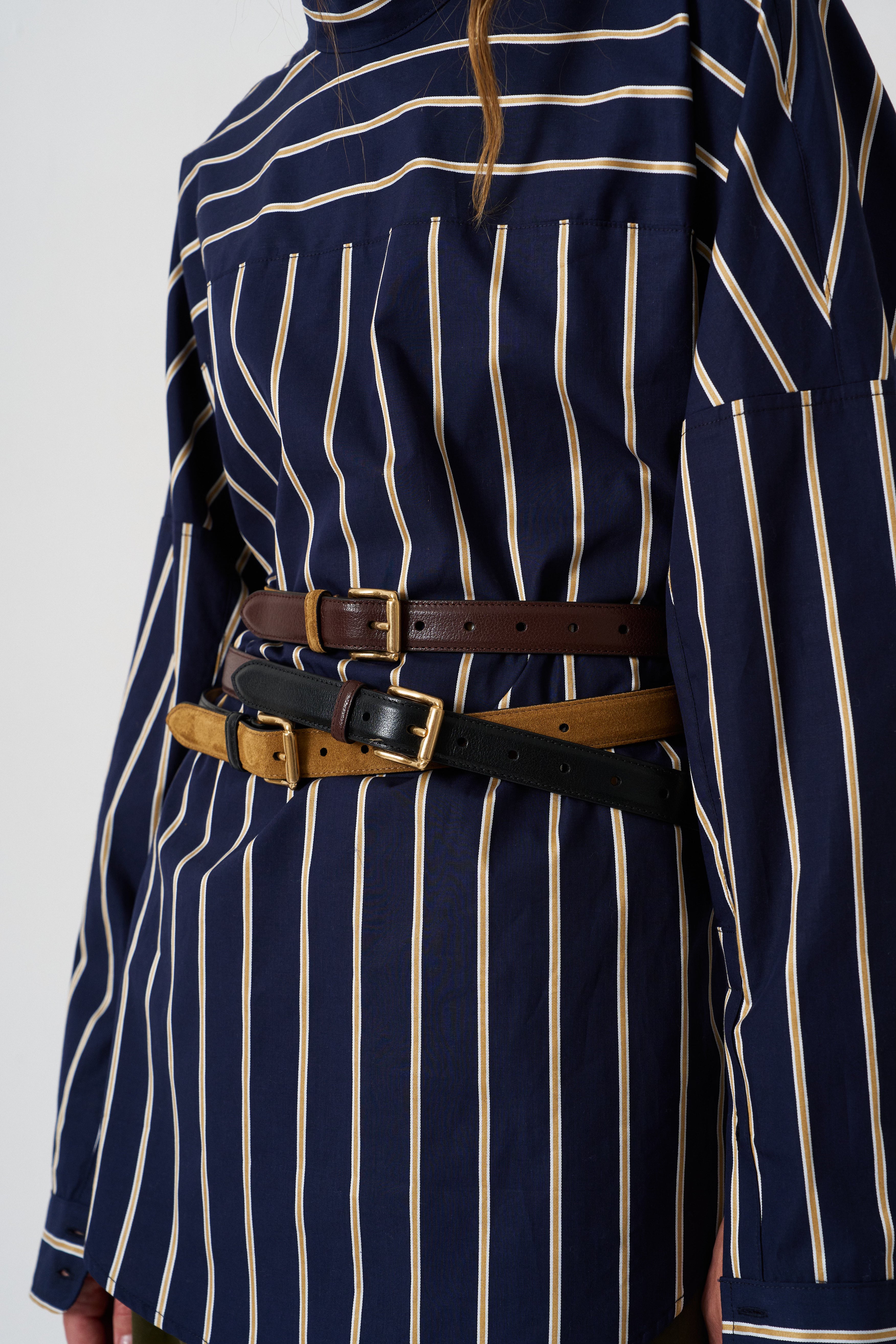 A close-up of a navy blue shirt dress with vertical cream stripes, styled with three leather belts in brown, tan, and black, worn by a model.
