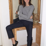 A woman sits on a wooden console table in front of a large gold-framed mirror, wearing a light gray knit sweater, dark navy cropped trousers, and black loafers.