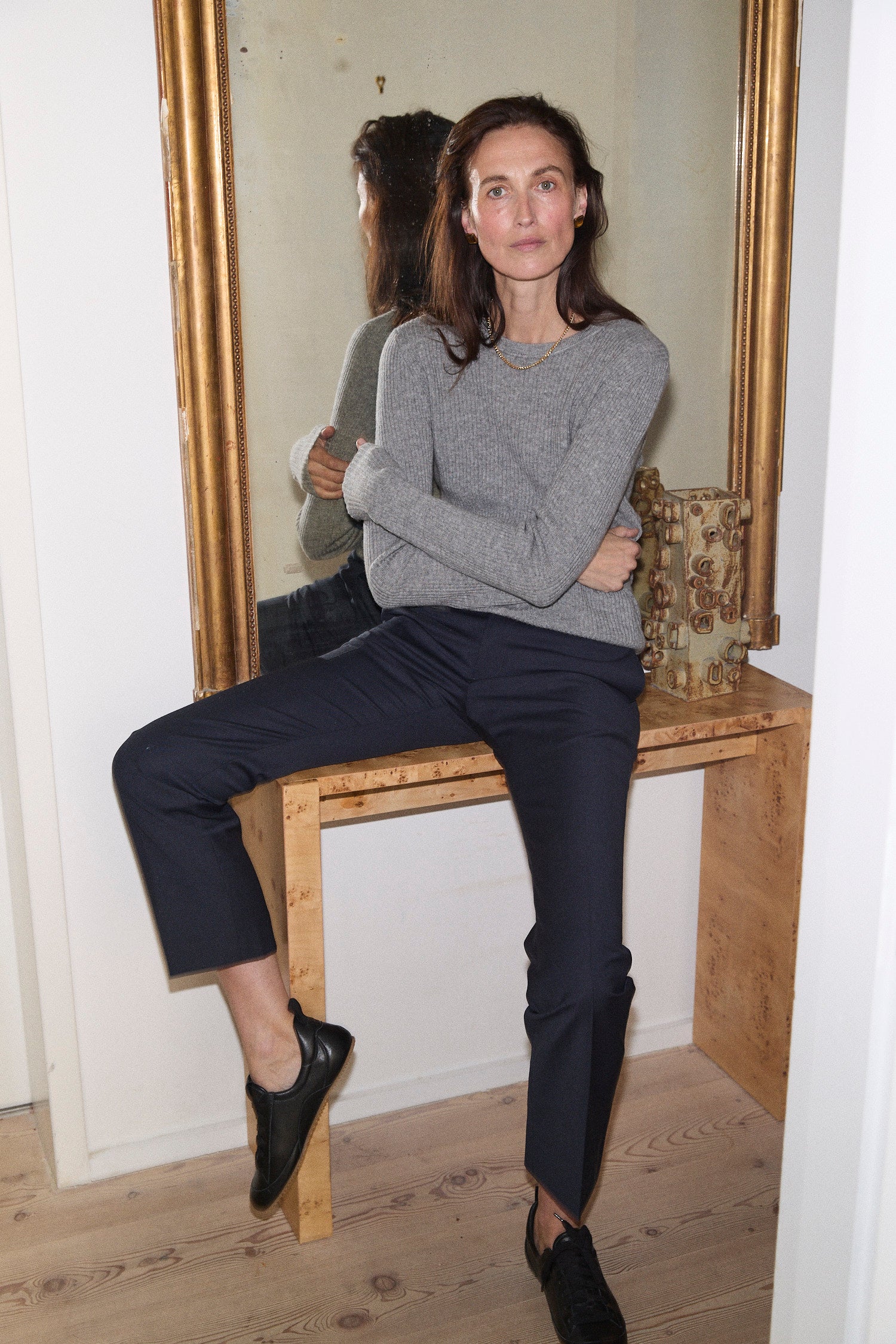 A woman sits on a wooden console table in front of a large gold-framed mirror, wearing a light gray knit sweater, dark navy cropped trousers, and black loafers.