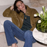 A woman sits on a light beige sofa wearing an olive brown leather jacket, a dark green top, and blue jeans. A glass vase of white tulips sits on a marble side table next to her.