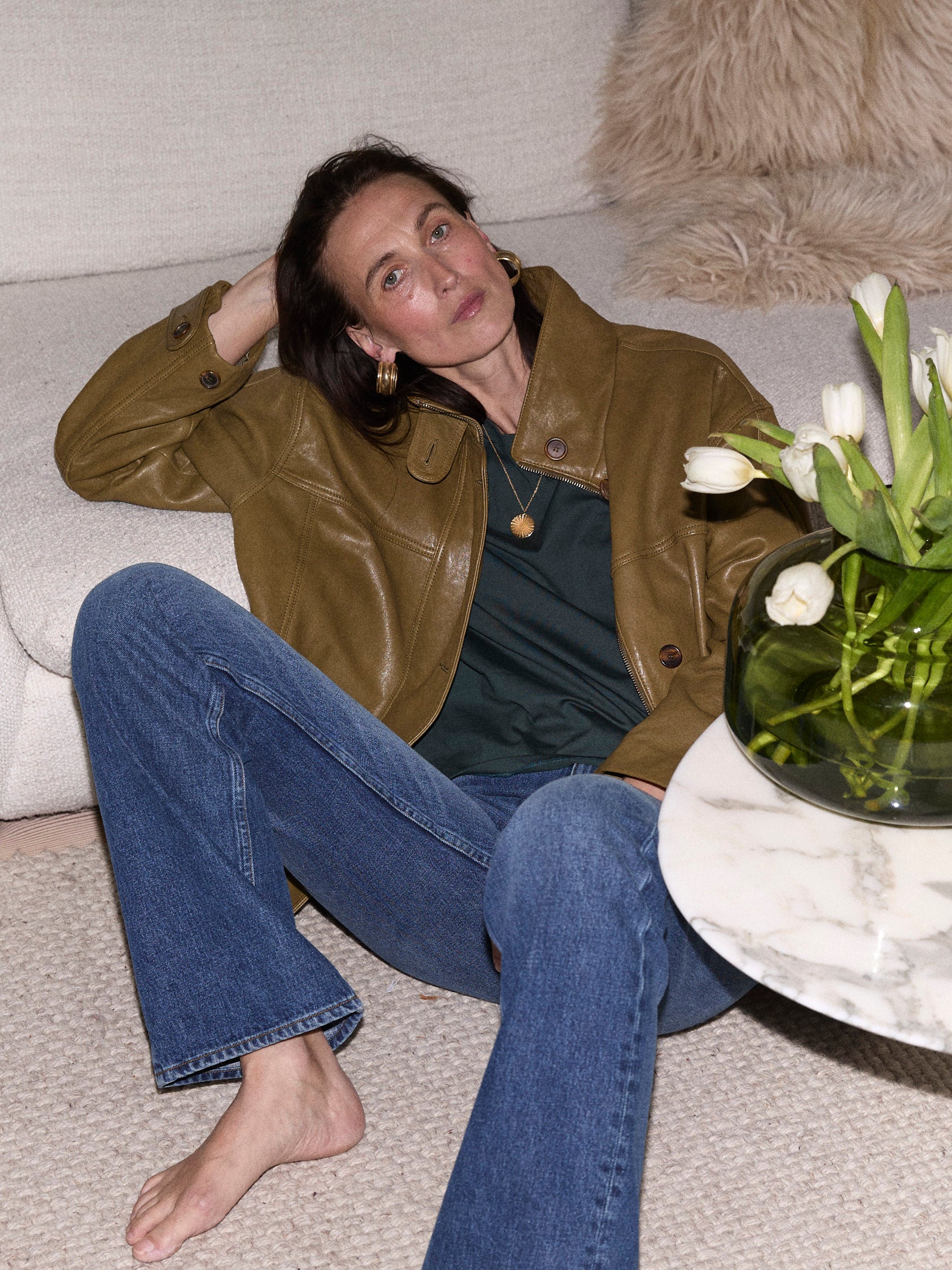 A woman sits on a light beige sofa wearing an olive brown leather jacket, a dark green top, and blue jeans. A glass vase of white tulips sits on a marble side table next to her.