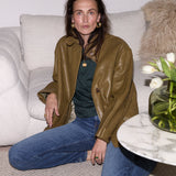 A woman sits on a light beige sofa wearing an olive brown leather jacket over a dark green top and blue jeans. A small marble side table with white tulips is in the foreground.