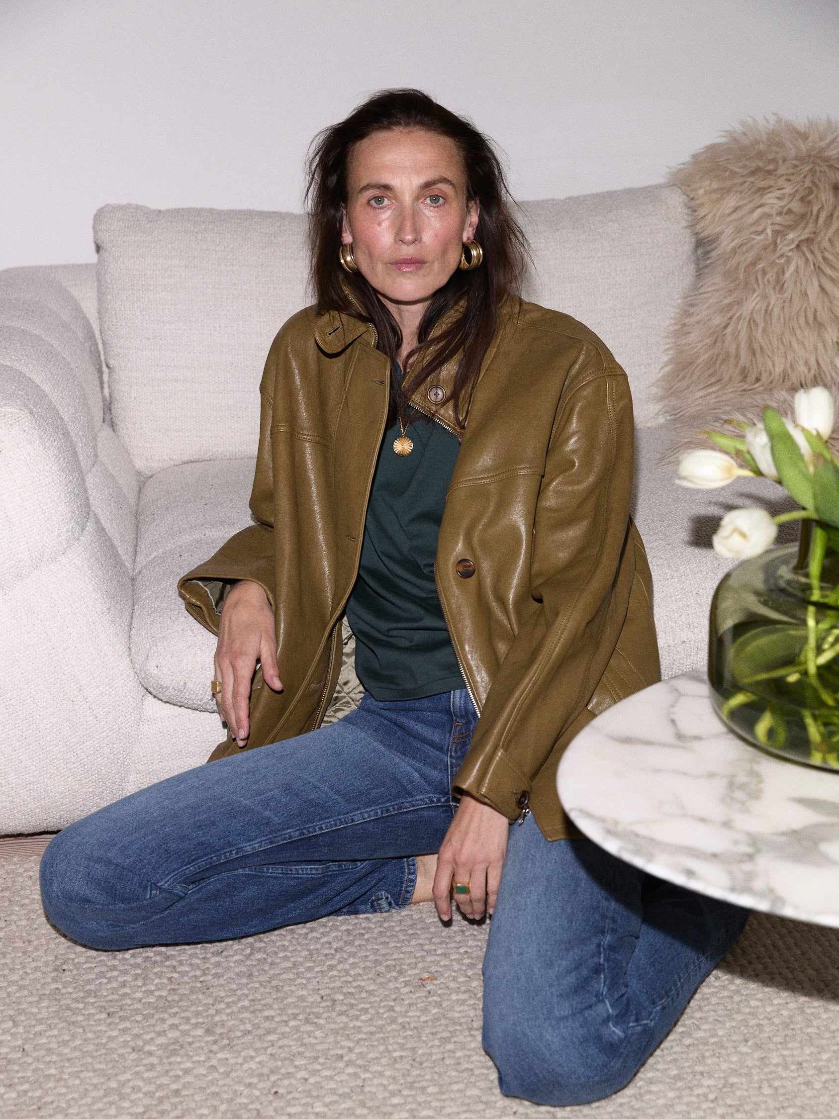 A woman sits on a light beige sofa wearing an olive brown leather jacket over a dark green top and blue jeans. A small marble side table with white tulips is in the foreground.