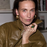 A woman with dark hair pulled back, wearing a shiny olive-brown leather jacket and large gold hoop earrings, posing indoors with a bookshelf blurred in the background.