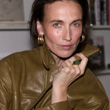 A woman with dark hair pulled back, wearing a shiny olive-brown leather jacket and large gold hoop earrings, posing indoors with a bookshelf blurred in the background.