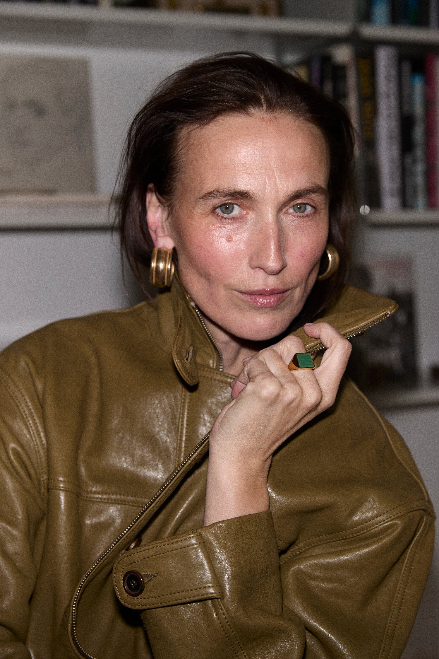 A woman with dark hair pulled back, wearing a shiny olive-brown leather jacket and large gold hoop earrings, posing indoors with a bookshelf blurred in the background.