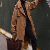 A woman stands on a city street beside a tall dark green construction wall, wearing a camel trench coat over a black outfit and holding a dark red leather tote bag.