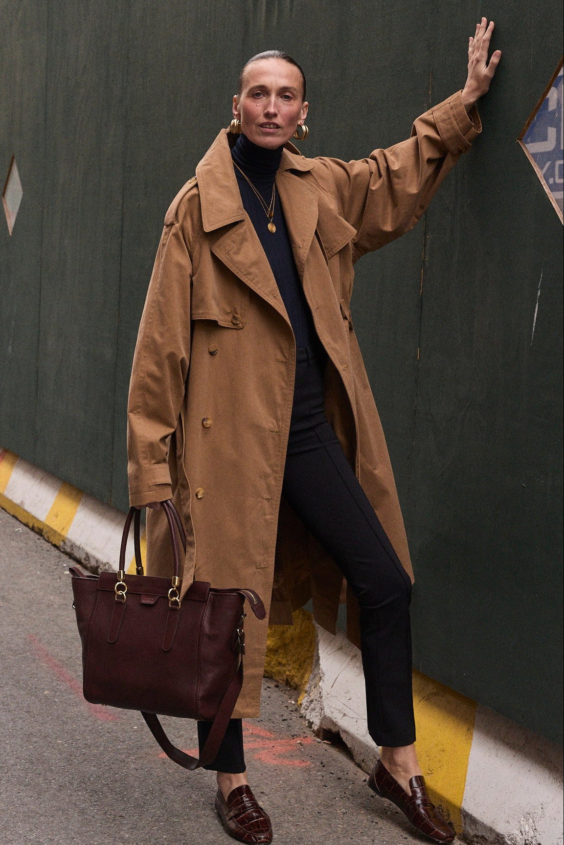 A woman stands on a city street beside a tall dark green construction wall, wearing a camel trench coat over a black outfit and holding a dark red leather tote bag.
