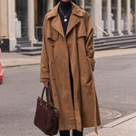 A woman stands on a city street wearing a long camel trench coat over a black turtleneck and black pants, holding a dark brown leather tote bag. Historic stone buildings with columns are in the background.