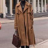 A woman stands on a city street wearing a long camel trench coat over a black turtleneck and black pants, holding a dark brown leather tote bag. Historic stone buildings with columns are in the background.