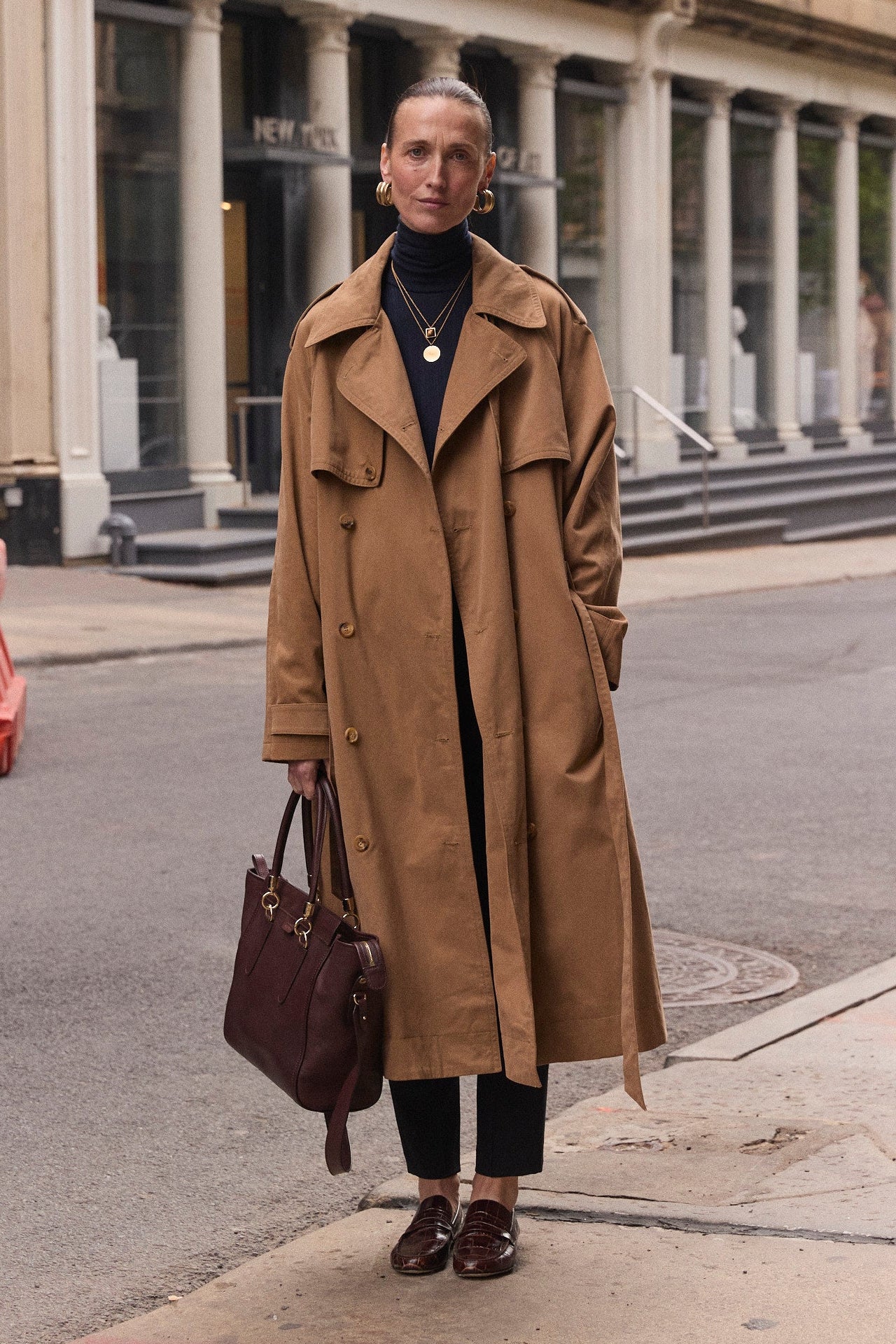 A woman stands on a city street wearing a long camel trench coat over a black turtleneck and black pants, holding a dark brown leather tote bag. Historic stone buildings with columns are in the background.