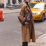 A woman stands on a city street wearing a long camel trench coat over a black turtleneck and black trousers, carrying a large dark brown leather shoulder bag. A yellow taxi is visible behind her.