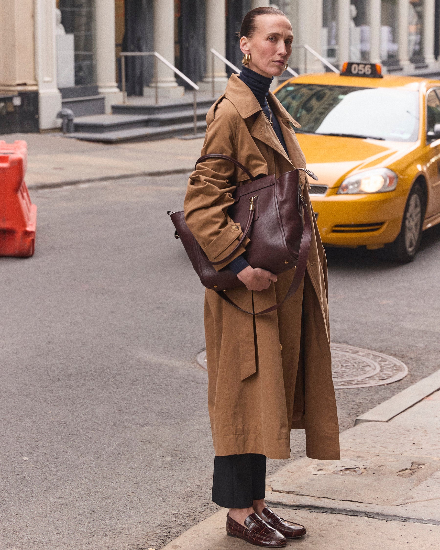 A woman stands on a city street wearing a long camel trench coat over a black turtleneck and black trousers, carrying a large dark brown leather shoulder bag. A yellow taxi is visible behind her.