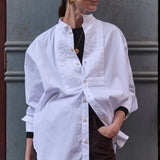 woman leaning against a grey wall in white shirt and brown pants