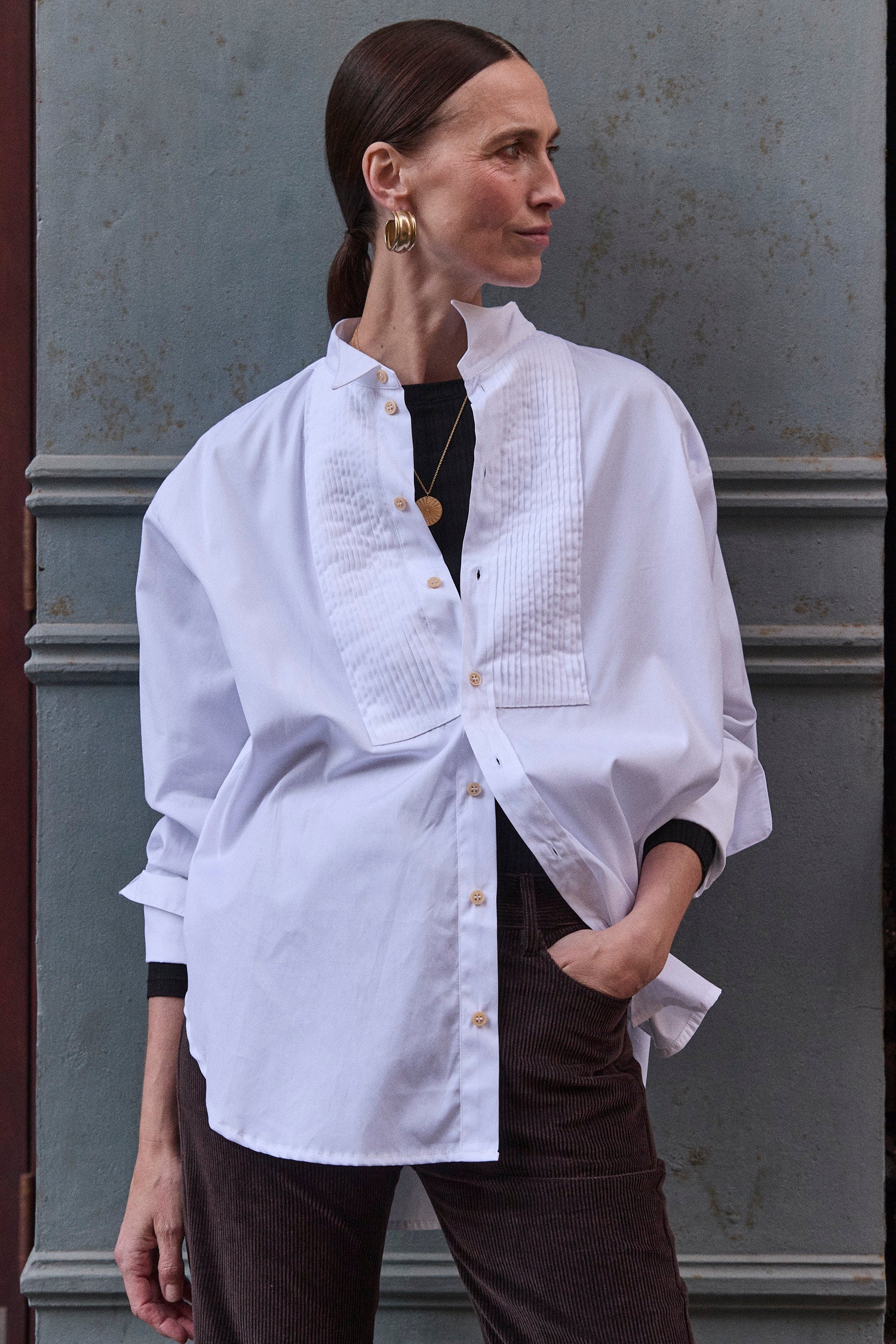 woman leaning against a grey wall in white shirt and brown pants