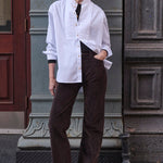 woman leaning against a grey wall in white shirt and brown pants