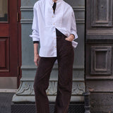 woman leaning against a grey wall in white shirt and brown pants