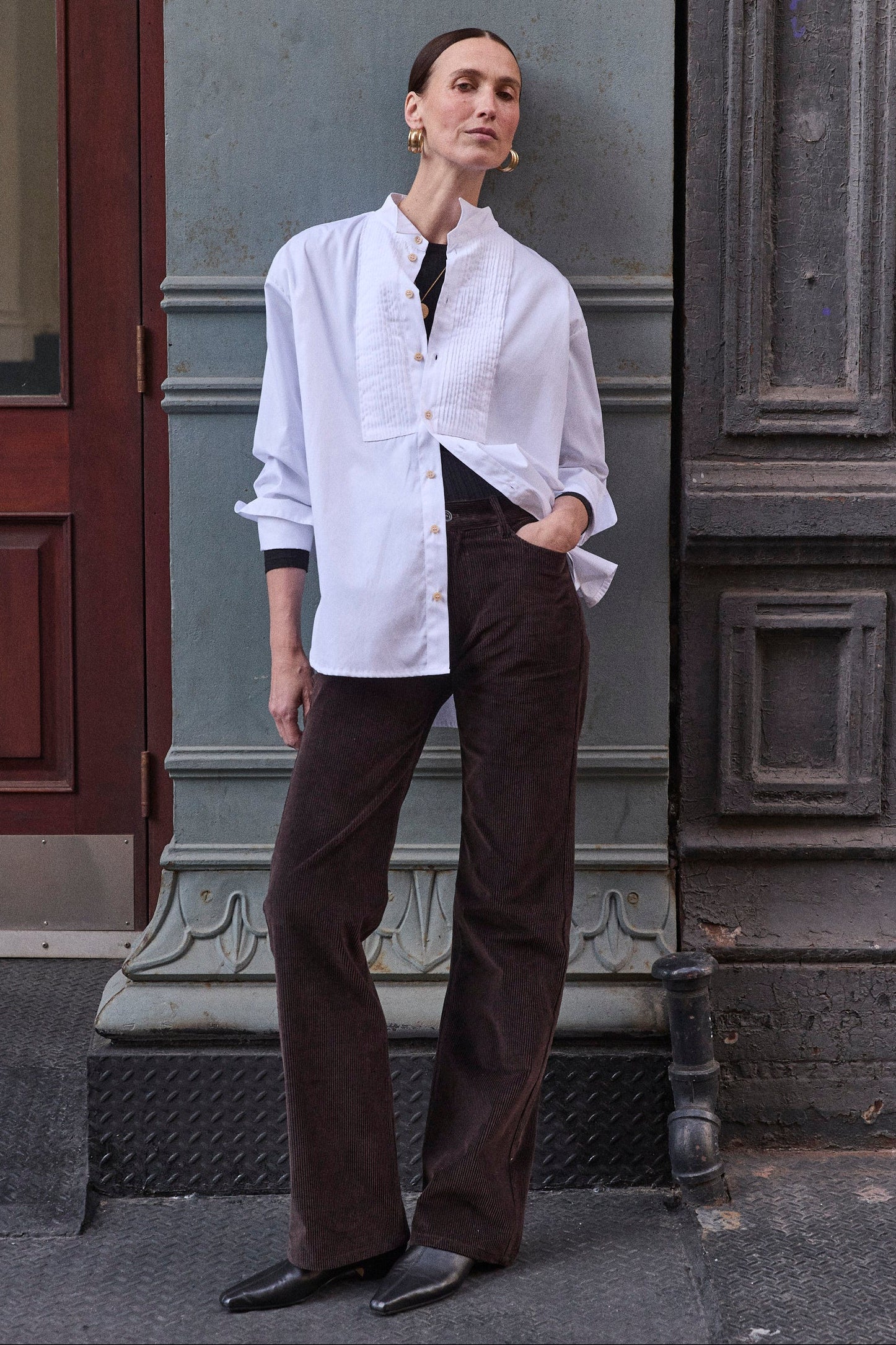 woman leaning against a grey wall in white shirt and brown pants